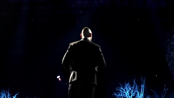 Man entertainer, presenter or actor on stage. Back, arms to sides, smoke on background of spotlight. Rear view of a male public speaker speaking at the microphone, pointing, in the — Stock Video