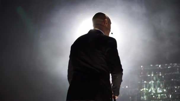 Man entertainer, presenter or actor on stage. Back, arms to sides, smoke on background of spotlight. Rear view of a male public speaker speaking at the microphone, pointing, in the — Stock Video