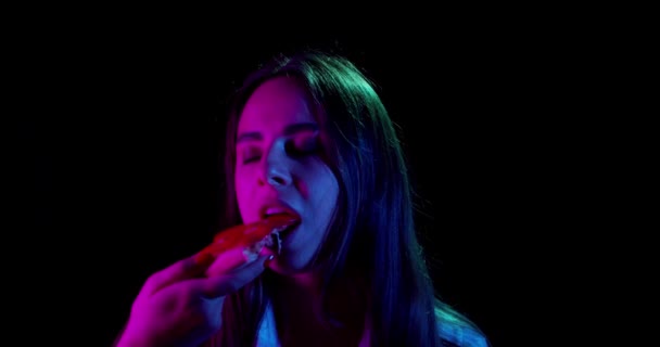 Gyönyörű lány eszik pizza neon fények egy fekete háttér — Stock videók