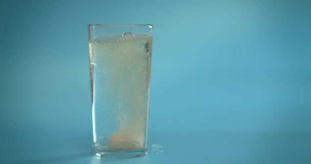 Een vitamine C tablet wordt met water in een doorzichtig glas gegooid. De bruisende roze tablet lost op. Glas op een blauwe achtergrond. Langzame beweging — Stockvideo