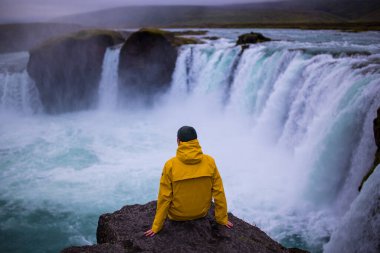 Sarı ceketli genç yürüyüşçü İzlanda 'daki Godafoss şelalesinde oturuyor. Godafoss, İzlanda 'da tanrıların şelalesi anlamına gelir..
