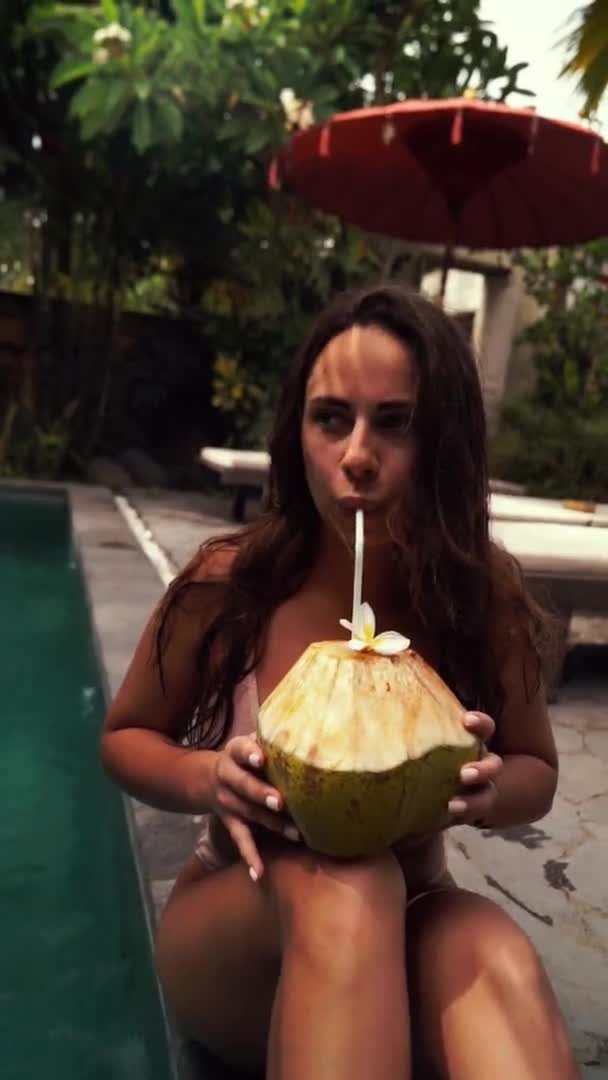 La joven hermosa mujer en bikini bebe leche de coco de un coco en el borde de la piscina en el complejo tropical. Vídeo vertical — Vídeos de Stock