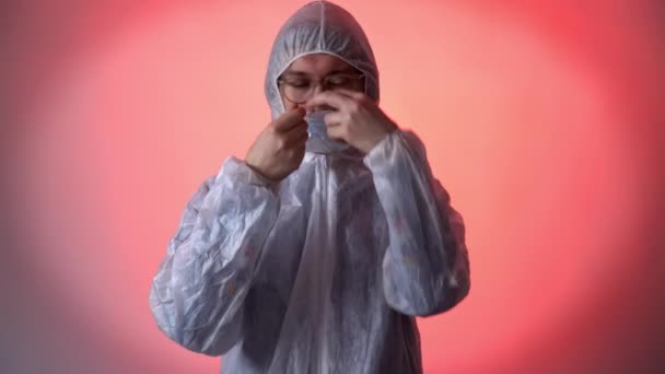 Kırmızı arka planda beyaz koruyucu giysili ciddi bir adam ilaç maskesi, fermuar ceketi ve kapüşon takıyor. Coronavirus covid-19 konsepti — Stok video