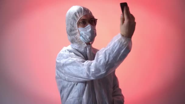 Человек в защитном костюме, медицинской маске и очках делает селфи на телефоне на красном фоне — стоковое видео