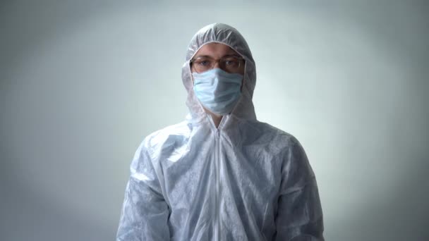 Homem sério em um terno protetor, máscara médica e óculos mostra um coração de rosas no fundo branco — Vídeo de Stock