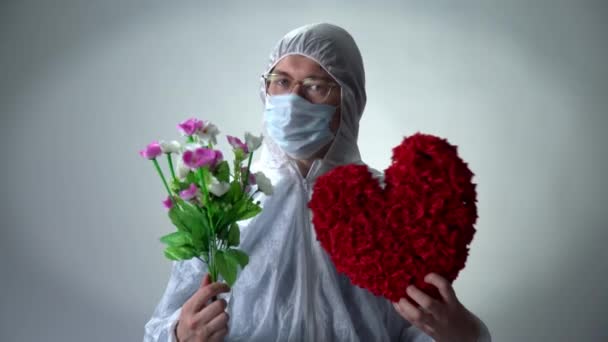 Een man in een beschermend pak, medisch masker en bril toont een hart van rozen en bloemen op een witte achtergrond — Stockvideo