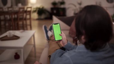 Ev soyutlanmış. Evdeki adam kanepede yatarken Smartphone 'u yeşil ekranla kullanıyor, Swiping yapıyor, Jestler yapıyor. Cep Telefonu Kullanan Adam, İnternet Sosyal Ağ Taraması.
