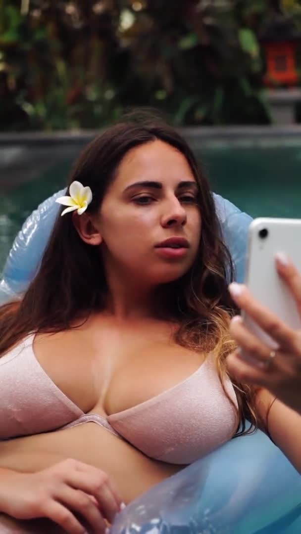 A beautiful woman is relaxing in the pool at a luxury beach resort spa hotel taking a selfie on her phone. Vertical video — Stock Video