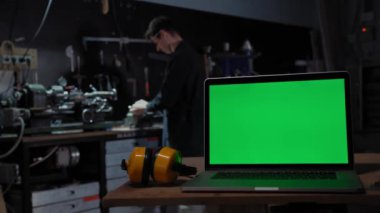 Endüstriyel çevrenin arka planında yeşil ekranlı bir laptop. Bir işçi bir değirmen makinesi için çalışır. Üretim ve inşaat kavramı