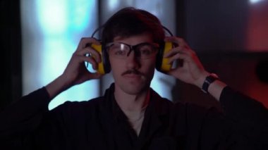 Siyah gömlekli, koruyucu gözlüklü, bıyıklı adam atölyede duruyor ve kulaklıkları takıyor..