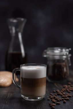 Şeffaf kahve kapuçinosu veya latte, siyah masada kurabiyeler ve bir kavanoz kahve çekirdeğinin yanında. Bir fincan kahve kapuçino ya da tarçınlı latte ve siyah ahşap kahve çekirdekleri.