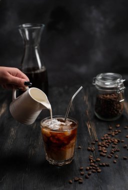 Bir kadın soğuk kahveye krema döker. Buzlu kahve, krema ve kahve çekirdekleri ile siyah taşlı arka planda.