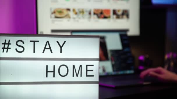 Ein Schild mit dem Text "Bleib zu Hause", eine Person, die am Computer arbeitet, im Neonlicht auf dem Hintergrund. COVID-19 Coronavirus STAY HOME SAVE LIVES virales Social-Media-Nachrichtenschild mit Text für soziale Netzwerke — Stockvideo