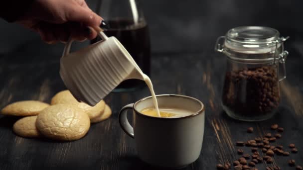 Повільний рух вершків вливається в чашку кави на чорному дерев'яному столі з кавовими зернами та печивом на чорному тлі — стокове відео