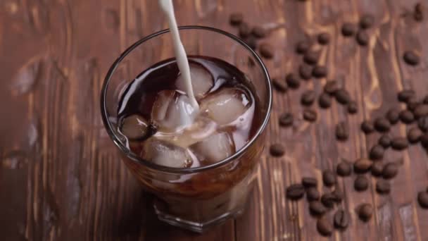 Tampilan teratas Gerakan krim yang dituangkan ke dalam segelas es kopi dingin di atas meja kayu cokelat dengan biji kopi dan kue di latar belakang hitam — Stok Video