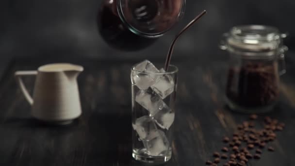 Add black coffee of cream being poured into a glass of cold brew iced coffee on black wood table with coffee beans on black background — Stock Video