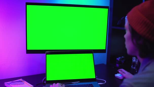 Knappe freelancer man fotograaf die naar laptop kijkt, typt op toetsenbord Twee monitoren met groen scherm in neon licht. Middelgroot Film Camera op tafel. Home zelf-isolatie quarantaine coronavirus — Stockvideo