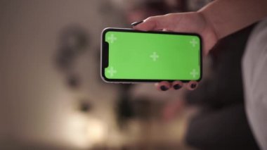 Ev soyutlanmış. Arka planda, elinde krom renkli yeşil ekran akıllı telefon tutan bir kadın içeriği dokunmadan ya da aşırmadan izliyor. Coronavirus Covid-19 uzaktan eğitim ya da çalışma konsepti karantinası. Yakın plan.
