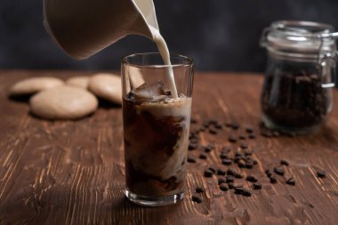 Kahverengi ahşap sehpanın üzerinde, kahve çekirdekleri ve siyah arka planda kurabiyeler olan uzun bir bardak buzlu kahveye krema dökülüyor.