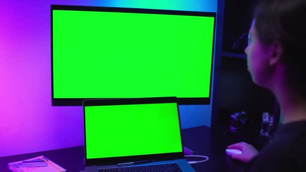 Mulher freelancer bonito olhando para laptop, trabalhar em casa on-line, digitando no teclado Dois monitores com tela verde em luz de néon. Câmera de filme de médio formato na mesa. Quarentena de auto-isolamento em casa — Vídeo de Stock