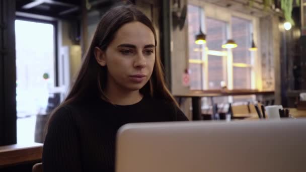 Joven hermosa morena trabaja en un ordenador portátil en Cool Creative Agency en una oficina Loft. Ella tiene un café para llevar y un cuaderno en su mesa. Cámara se acerca y ella sonríe y ríe. — Vídeos de Stock