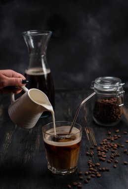 Bir kadın kahveye krema döker. Kremalı buzlu kahve bardağı ve siyah taşlı kahve çekirdeği.