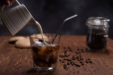 Bir kadın soğuk kahveye krema döker. Kahverengi ahşap masada sütlü buzlu kahve, kahve çekirdekleri ve siyah arka planda kurabiyeler.