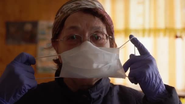 Літня жінка одягає захисну медичну маску біля вікна, дивиться на камеру. Профілактика пандемії коронавірусу COVID-19. Соціальне дистанціювання — стокове відео