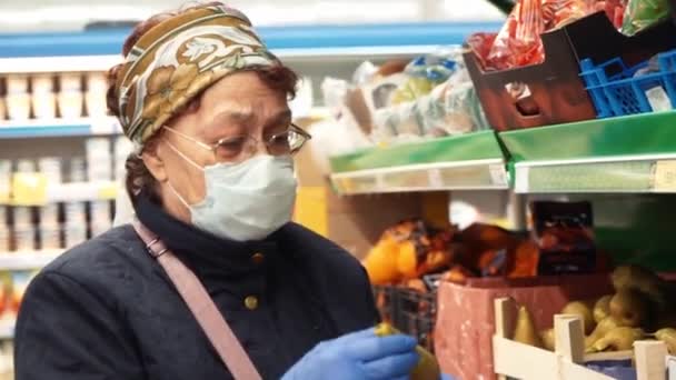Oudere vrouw in beschermende medische masker en geneeskunde handschoenen kiezen producten in de supermarkt. De coronavirusepidemie in de stad moet thuis blijven. COVID-19 Pandemie. Sociale afstand — Stockvideo