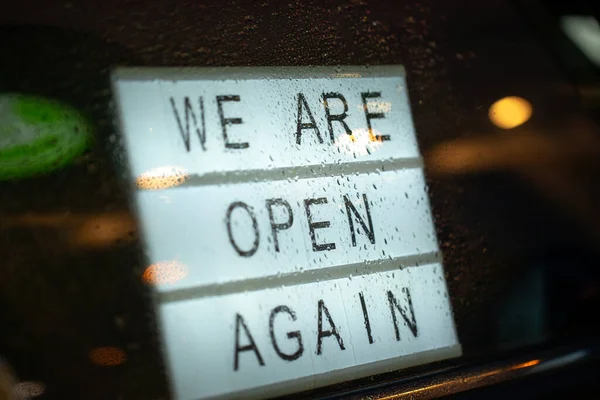 Lightbox with a sign we are open again behind a glass door of the cafe during the rain in the evening. Were open again after quarantine, photo of small business owner. Please wear a face mask and — Stock Photo, Image