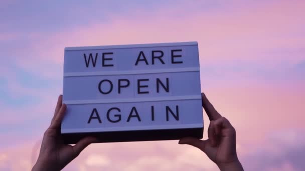 Handen houden een Lightbox vast met een teken We Are Open Again, roze blauwe lucht op de achtergrond. Waren weer open na quarantaine, video van kleine ondernemer eigenaar. COVID-19 pandemische preventie van coronavirus. — Stockvideo