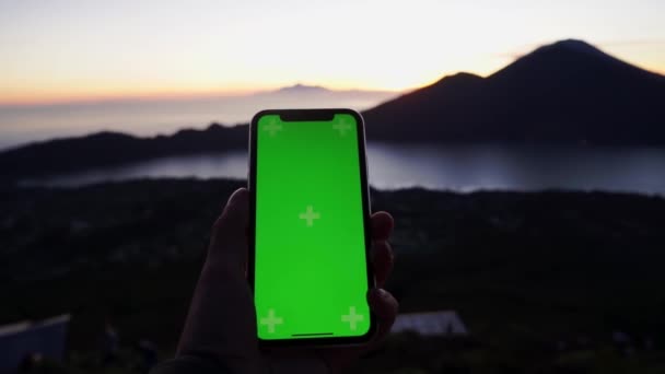 스마트폰을 들고 있는 남성의 팔은 경치가 아름다운 곳에서 수직 위치에 있습니다. 가까이 서 구경하는 사람들은 산의 멋진 풍경을 보여 주는 초록빛 전화기와 함께 손을 잡는다. 선셋. 여행. — 비디오