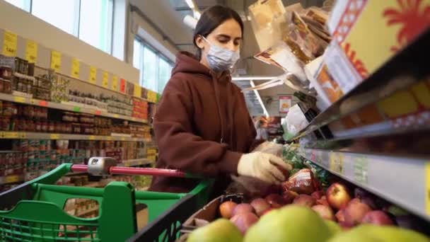 Красива жінка в медичній масці і рукавички ретельно вибирає яблука в продуктовому супермаркеті. Захист від коронавірусної епідемії, підвищений імунітет зі свіжими фруктами. Здорова їжа для боротьби — стокове відео