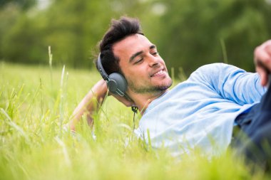 Yeşil çimenlerin üzerinde yatan sevimli genç adam müzik dinlemek ve bir tarafına bak