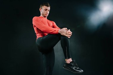 Kırmızı spor giyim giymek ve koyu arka plan ağır bir çalışmadan sonra onun bacak germe sportif genç adam. Güçlü yakışıklı atletik erkek uygun yapıyor. Spor motivasyon. Sağlıklı yaşam tarzı.