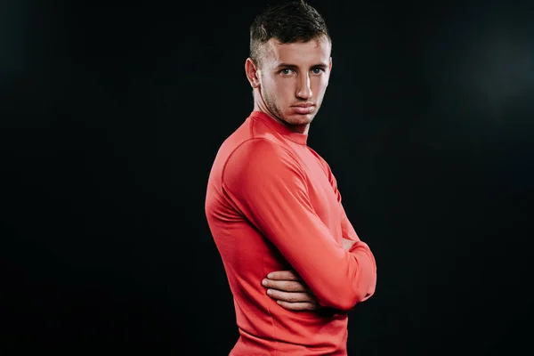 Profile view of attractive athlete man in red sportswear standing over dark background, thinking over problems in his life, holding hands, looking front. Lifestyle, sport, motivation.