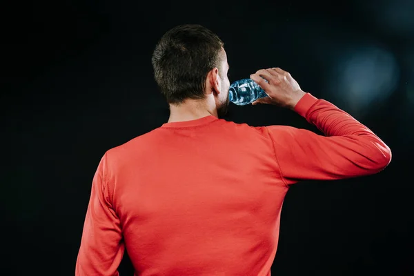 Rear shot portrait of handsome muscular Caucasian jogger wearing red sportswear drinking water, relaxing after intense physical training in gym. Sport motivation. Lifestyle.