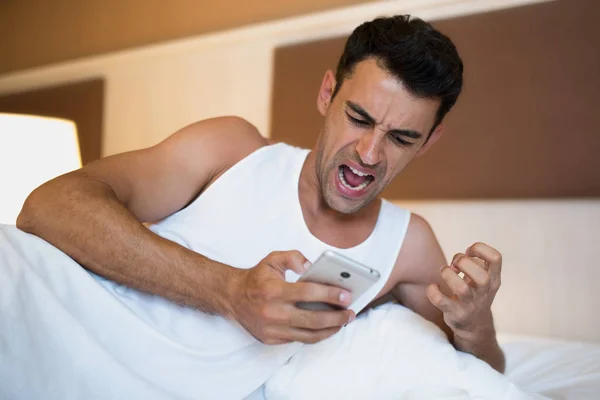 Guapo joven molesto usando un teléfono móvil en la cama blanca . — Foto de Stock