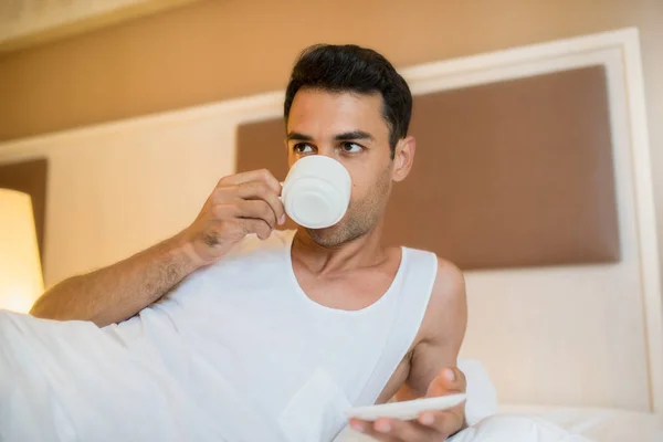 Guapo joven despierto bebiendo taza de café en la cama blanca — Foto de Stock