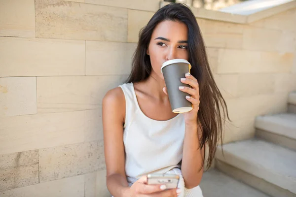 Belo retrato de jovem morena atraente bebendo com uma xícara de café ou chá, sentado nos degraus do parque e mensagens de texto no telefone celular. Olhando para um lado e pensando . — Fotografia de Stock