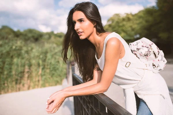 Mujer joven atractiva y hermosa con mochila apoyada en la barandilla y mira hacia adelante, en el fondo de la naturaleza en el parque. Viajar. Vacaciones —  Fotos de Stock