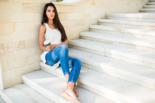 Retrato de corpo inteiro de bela mulher morena sentada nos degraus de um parque, no fundo do lago. Sorrindo com uma xícara de café ou chá. Olhando para a câmera . — Fotografia de Stock