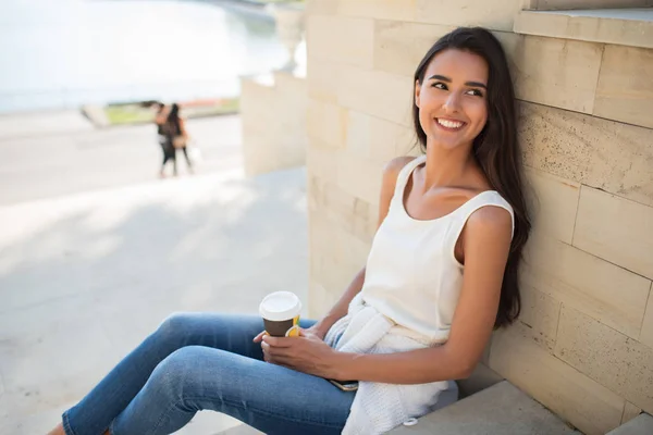Happy beautiful woman sitting on the steps of a park, on lake background. Smiling with a cup of coffee or tea. Looking at one side. — Stock Photo, Image