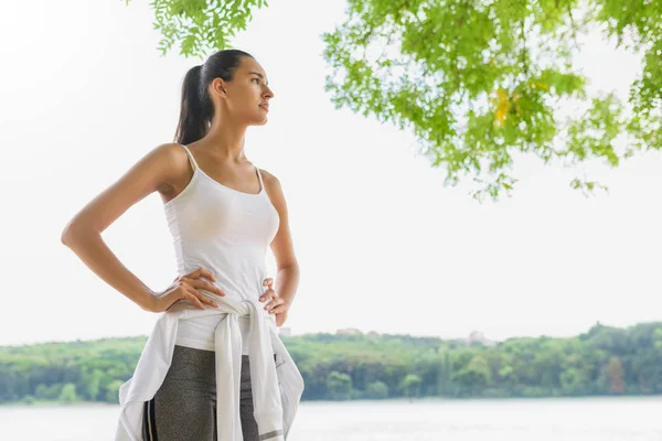 Bottom view of beautiful brunette female runner relax after running in city park with earphones, look at one side. Healthy fitness woman jogging outdoors. Nature background. Motivation sport. — Stock Photo, Image