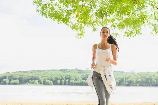 Mooie brunette vrouwelijke loper uitgevoerd in stadspark met koptelefoon. Gezonde fitness vrouw joggen buiten. Achtergrond van de natuur. Motivatie sport. — Stockfoto