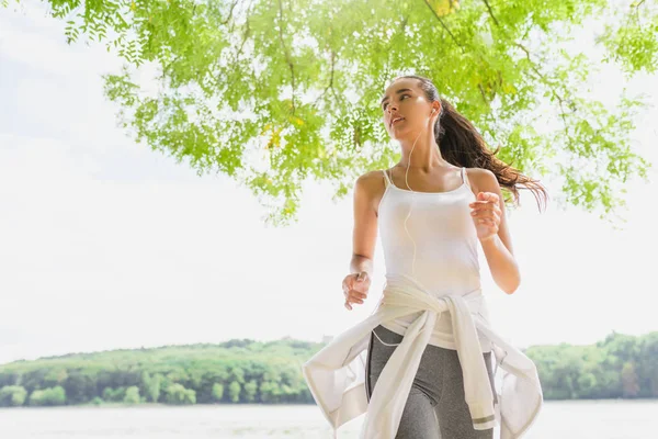 Vista inferiore di bella bruna corridore femminile in esecuzione nel parco della città con auricolari. Donna sana di fitness che corre all'aperto. Sfondo naturale. Sport motivazionale . — Foto Stock