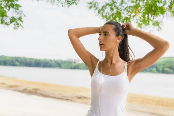 Attractive brunette young fitness woman in sportswear listening music with earphones after training outdoors at beautiful sunny day. Runner girl corrects tail and looking away. — Stock Photo, Image