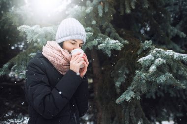 Güzel beyaz kadın kış ceket, bir fincan kahve ya da çay, açık havada park veya orman kış günü kar yağışı üzerinde tutarak gri bere şapka ve pembe eşarp, ördü. Kış ruh hali.