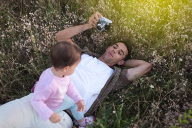 Genç şirin mutlu yürümeye başlayan çocuk kızının oynarken ve çim üzerinde gülüyor için yalancı yüzünü Peder. Mutlu baba onun bebek kız fotoğraf ile fotoğraf makinesi ile. Çayır üzerinde yürüme aile.