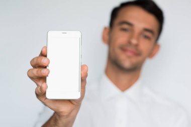 Yakışıklı delikanlı bir ışık gri arka plan üzerinde izole bir boş akıllı telefon ekranını göstermeyi portresi. Bulanık gülümseme beyaz işadamı ile telefon Studio.